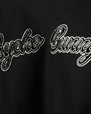 Close-up of a black fabric on the MENS ROBBINS VARSITY BOMBER JACKET (model B6J524Z1OW) by Psycho Bunny, showcasing the text "Psycho Bunny" in a cursive, embroidered style. The white text with a black outline contrasts against the wool-blend background, which is decorated with plush chenille patches.