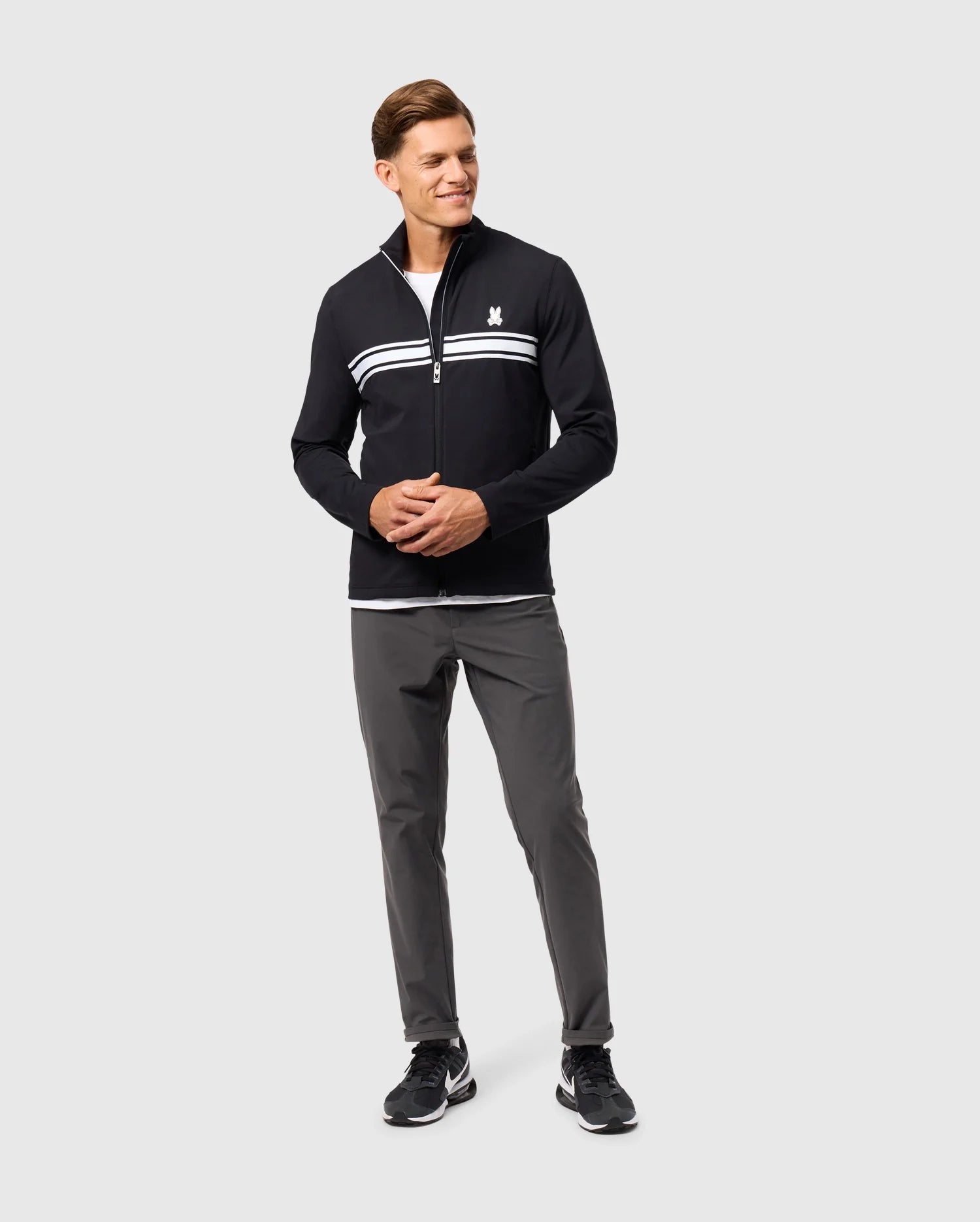 A man is standing against a plain background, wearing the MENS TERRELL SPORT ZIP UP JACKET - B6J643C200 by Psycho Bunny, complemented by gray pants and black sneakers. He is smiling and has his hands clasped in front.