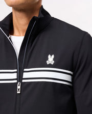 A close-up of a person wearing the Psycho Bunny MENS TERRELL SPORT ZIP UP JACKET – B6J643C200, which showcases white horizontal stripes and a small logo featuring a stylized rabbit above crossed bones on the chest. This sporty jacket includes a zipper with a small rabbit logo on the pull tab, seamlessly combining style and comfort.