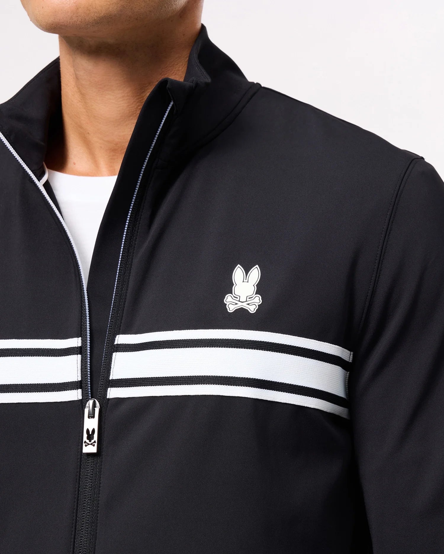 A close-up of a person wearing the Psycho Bunny MENS TERRELL SPORT ZIP UP JACKET – B6J643C200, which showcases white horizontal stripes and a small logo featuring a stylized rabbit above crossed bones on the chest. This sporty jacket includes a zipper with a small rabbit logo on the pull tab, seamlessly combining style and comfort.