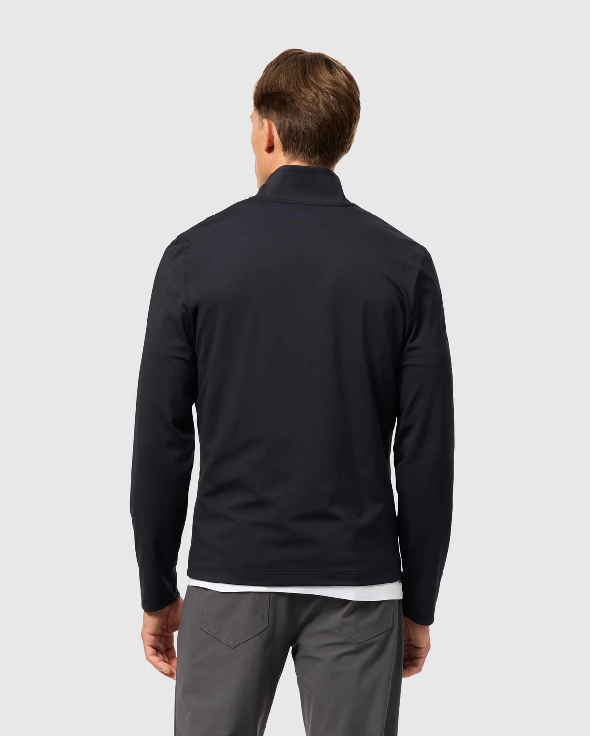 A person with short brown hair stands with their back to the viewer, wearing a sleek Psycho Bunny MENS TERRELL SPORT ZIP UP JACKET - B6J643C200 over a white shirt and gray pants. This versatile track jacket adds an element of style against the plain, light-colored background, blending effortlessly with its surroundings.