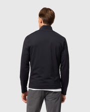 A person with short brown hair stands with their back to the viewer, wearing a sleek Psycho Bunny MENS TERRELL SPORT ZIP UP JACKET - B6J643C200 over a white shirt and gray pants. This versatile track jacket adds an element of style against the plain, light-colored background, blending effortlessly with its surroundings.