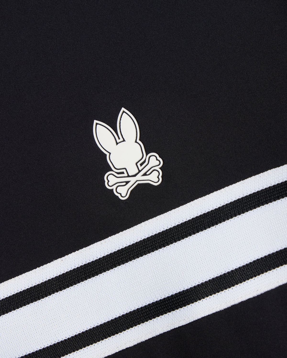 This Psycho Bunny MENS TERRELL SPORT ZIP UP JACKET - B6J643C200 is a versatile track jacket made from a modal-blend knit. It features black fabric with the signature white rabbit and crossbones logo, complemented by two diagonally running white and black striped bands beneath the emblem for a sporty flair.