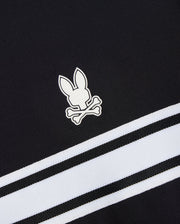 This Psycho Bunny MENS TERRELL SPORT ZIP UP JACKET - B6J643C200 is a versatile track jacket made from a modal-blend knit. It features black fabric with the signature white rabbit and crossbones logo, complemented by two diagonally running white and black striped bands beneath the emblem for a sporty flair.