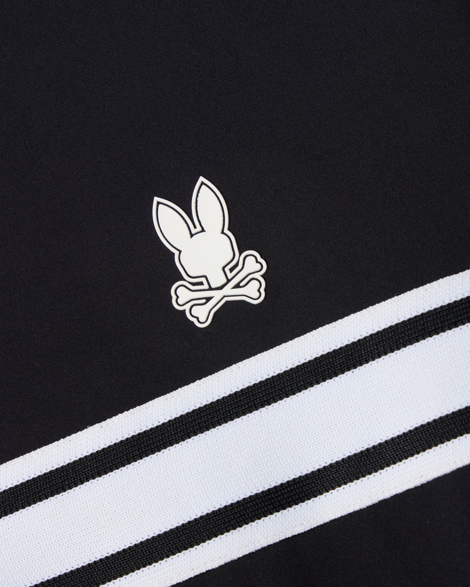 This Psycho Bunny MENS TERRELL SPORT ZIP UP JACKET - B6J643C200 is a versatile track jacket made from a modal-blend knit. It features black fabric with the signature white rabbit and crossbones logo, complemented by two diagonally running white and black striped bands beneath the emblem for a sporty flair.