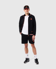 A person stands on a gray background wearing a black Psycho Bunny MENS COURTSIDE TRACK JACKET - B6J692C200 with orange details, a white polo shirt, black shorts with an orange logo, white socks, black athletic shoes, and a black visor with an orange emblem. They have hands in the jacket pockets, showcasing the comfort of stretch cotton-blend jersey.