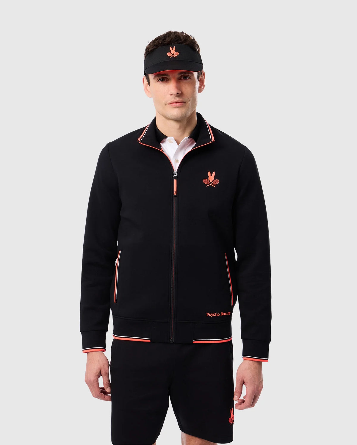 A person is standing against a plain background wearing a Psycho Bunny MENS COURTSIDE TRACK JACKET - B6J692C200 with red accents and a matching pair of shorts. They are also wearing a black visor. The stretch cotton-blend jersey jacket and shorts feature logo-engraved hardware and small red logos on the chest and left thigh, respectively.