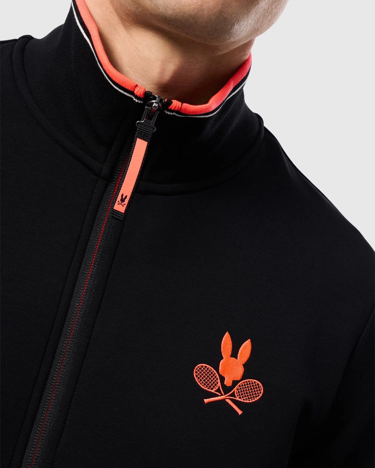 Close-up of a person wearing a black **Psycho Bunny MENS COURTSIDE TRACK JACKET - B6J692C200** in stretch cotton-blend jersey with a red zipper and an embroidered logo on the chest. The logo features a red rabbit head above two crossed tennis rackets. The collar lining is red with white accents. Only part of the face is visible.