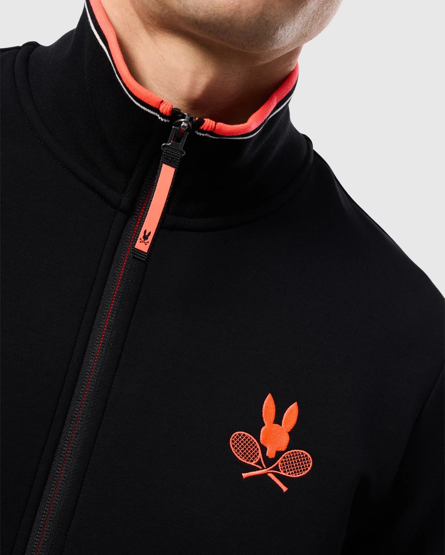 Close-up of a person wearing a black **Psycho Bunny MENS COURTSIDE TRACK JACKET - B6J692C200** in stretch cotton-blend jersey with a red zipper and an embroidered logo on the chest. The logo features a red rabbit head above two crossed tennis rackets. The collar lining is red with white accents. Only part of the face is visible.