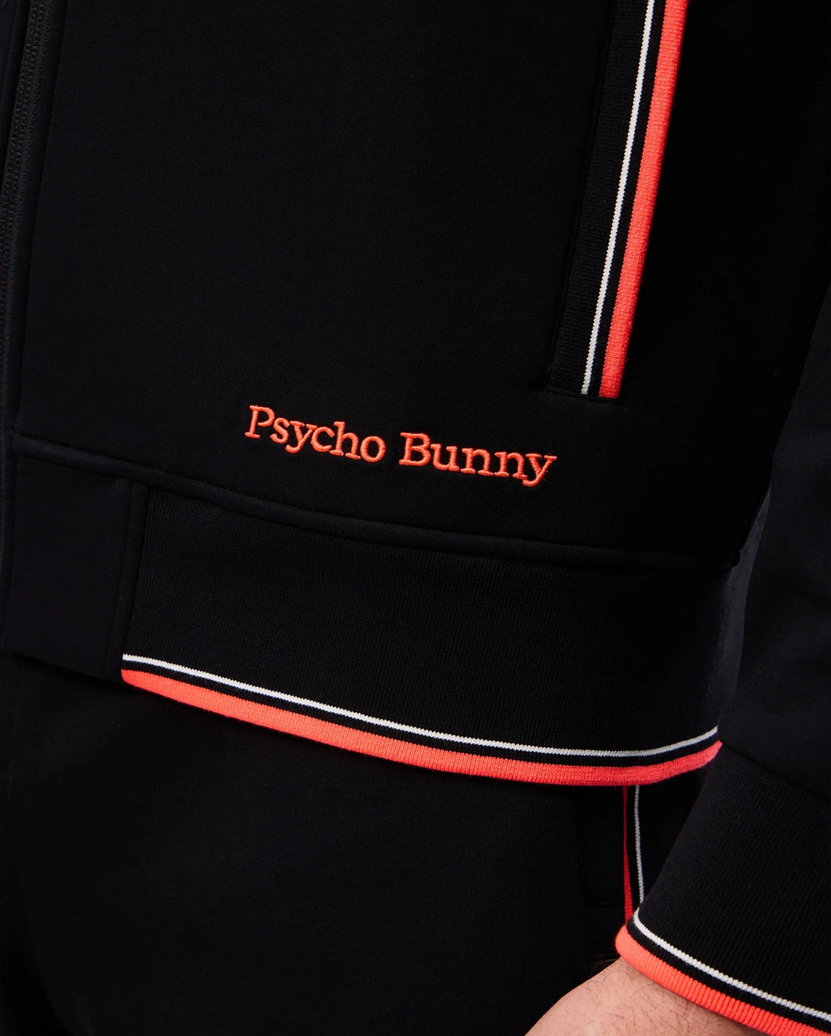 Close-up of a person wearing a black Psycho Bunny MENS COURTSIDE TRACK JACKET - B6J692C200 with “Psycho Bunny” embroidered in orange on the front. The jacket, made from stretch cotton-blend jersey, features orange and white striped trim on the zipper area and sleeve cuff, with logo-engraved hardware accents.