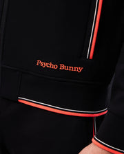 Close-up of a person wearing a black Psycho Bunny MENS COURTSIDE TRACK JACKET - B6J692C200 with “Psycho Bunny” embroidered in orange on the front. The jacket, made from stretch cotton-blend jersey, features orange and white striped trim on the zipper area and sleeve cuff, with logo-engraved hardware accents.