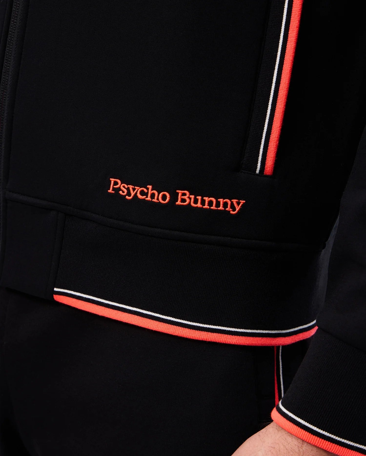 Close-up of a person wearing a black Psycho Bunny MENS COURTSIDE TRACK JACKET - B6J692C200 with “Psycho Bunny” embroidered in orange on the front. The jacket, made from stretch cotton-blend jersey, features orange and white striped trim on the zipper area and sleeve cuff, with logo-engraved hardware accents.