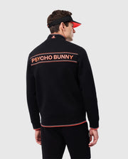 A man dressed in a black MENS COURTSIDE TRACK JACKET - B6J692C200 with orange trim and the words "Psycho Bunny" on the back. He is wearing a black visor with an orange brim. The background is plain and light gray.