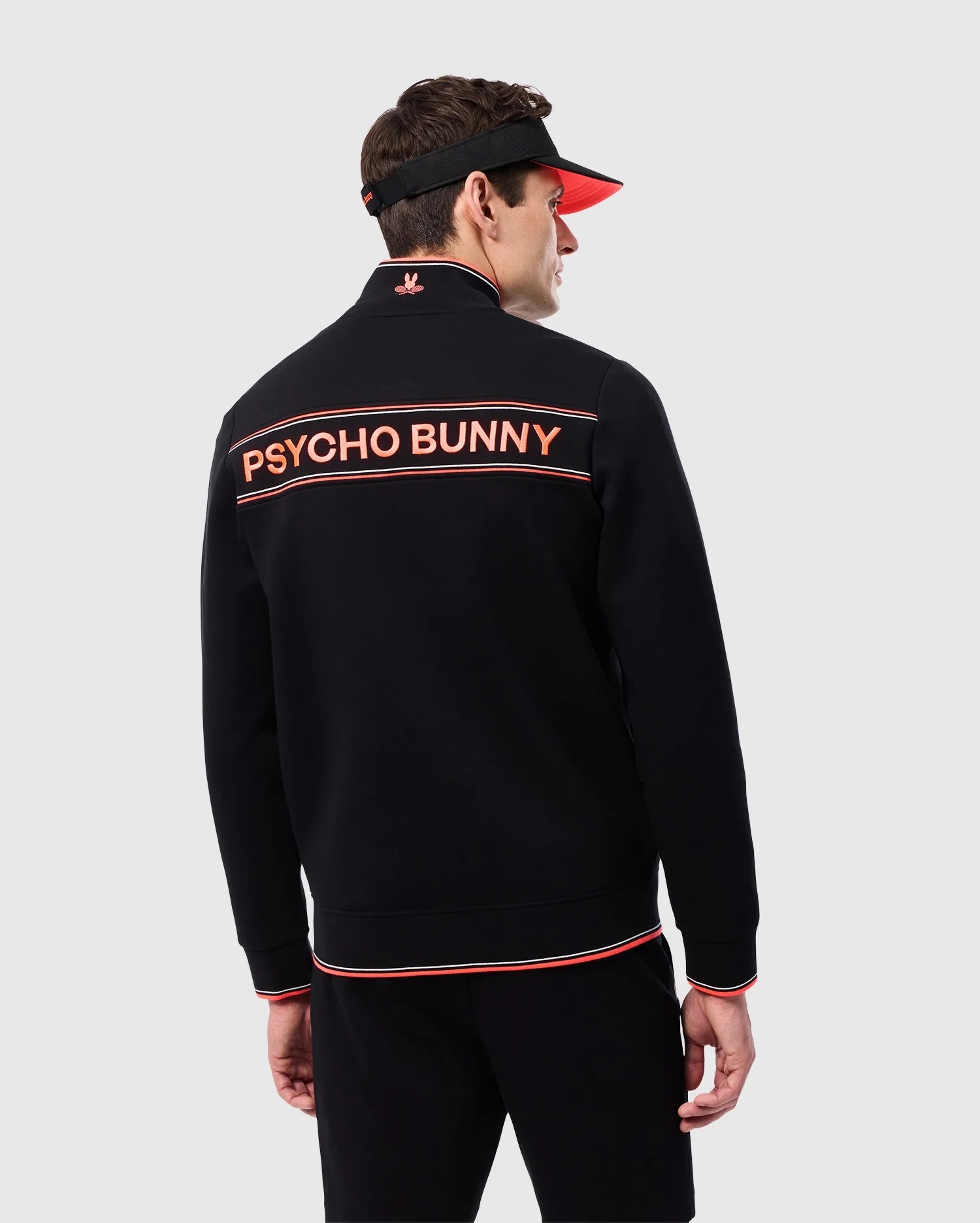 A man dressed in a black MENS COURTSIDE TRACK JACKET - B6J692C200 with orange trim and the words "Psycho Bunny" on the back. He is wearing a black visor with an orange brim. The background is plain and light gray.