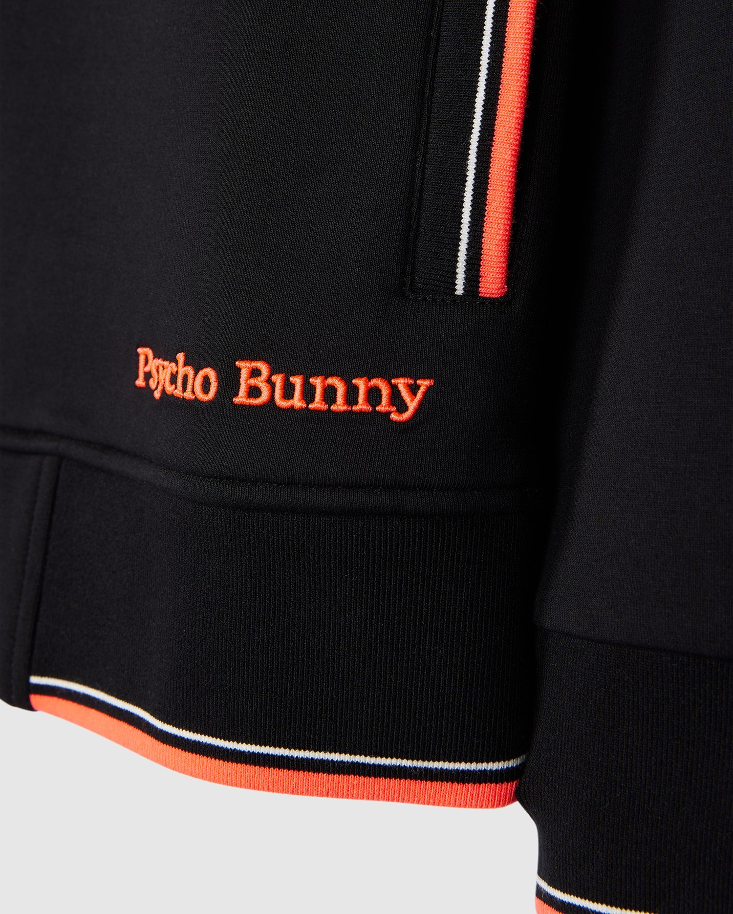 Close-up of a black Psycho Bunny MENS COURTSIDE TRACK JACKET - B6J692C200 with striped trim in orange, white, and black. The stretch cotton-blend jersey fabric features an embroidered "Psycho Bunny" logo in orange thread near the hem. The image highlights the texture, details of the stitching, and trim.