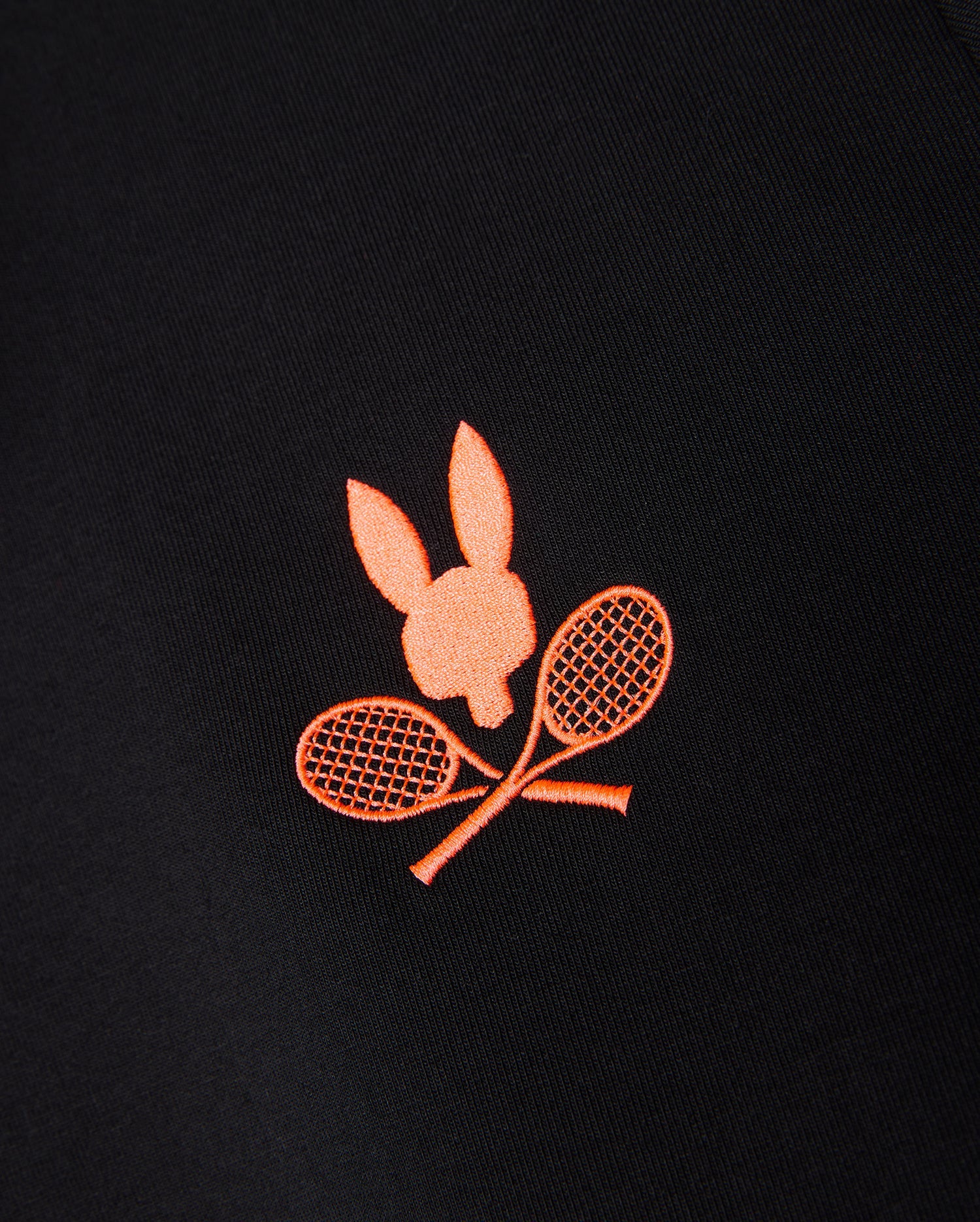 A close-up of a black Psycho Bunny MENS COURTSIDE TRACK JACKET - B6J692C200 featuring an orange embroidered design of a bunny head above two crossed tennis rackets.