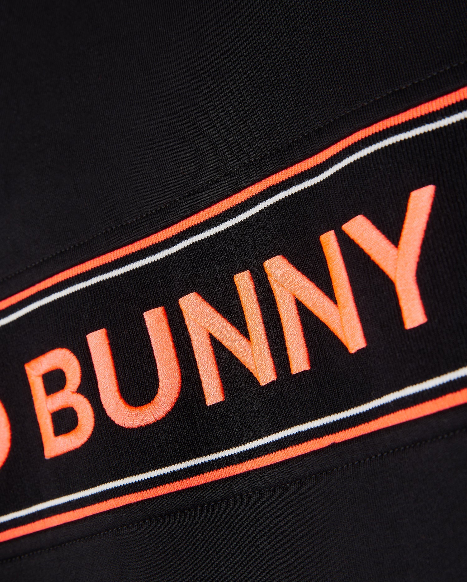 Close-up of a black **Psycho Bunny MENS COURTSIDE TRACK JACKET - B6J692C200** fabric with an orange and white stripe. The text "BUNNY" in bold orange letters is prominently displayed in the center of the stripe. The texture of the stretch cotton-blend jersey fabric is visible, giving it a detailed look.