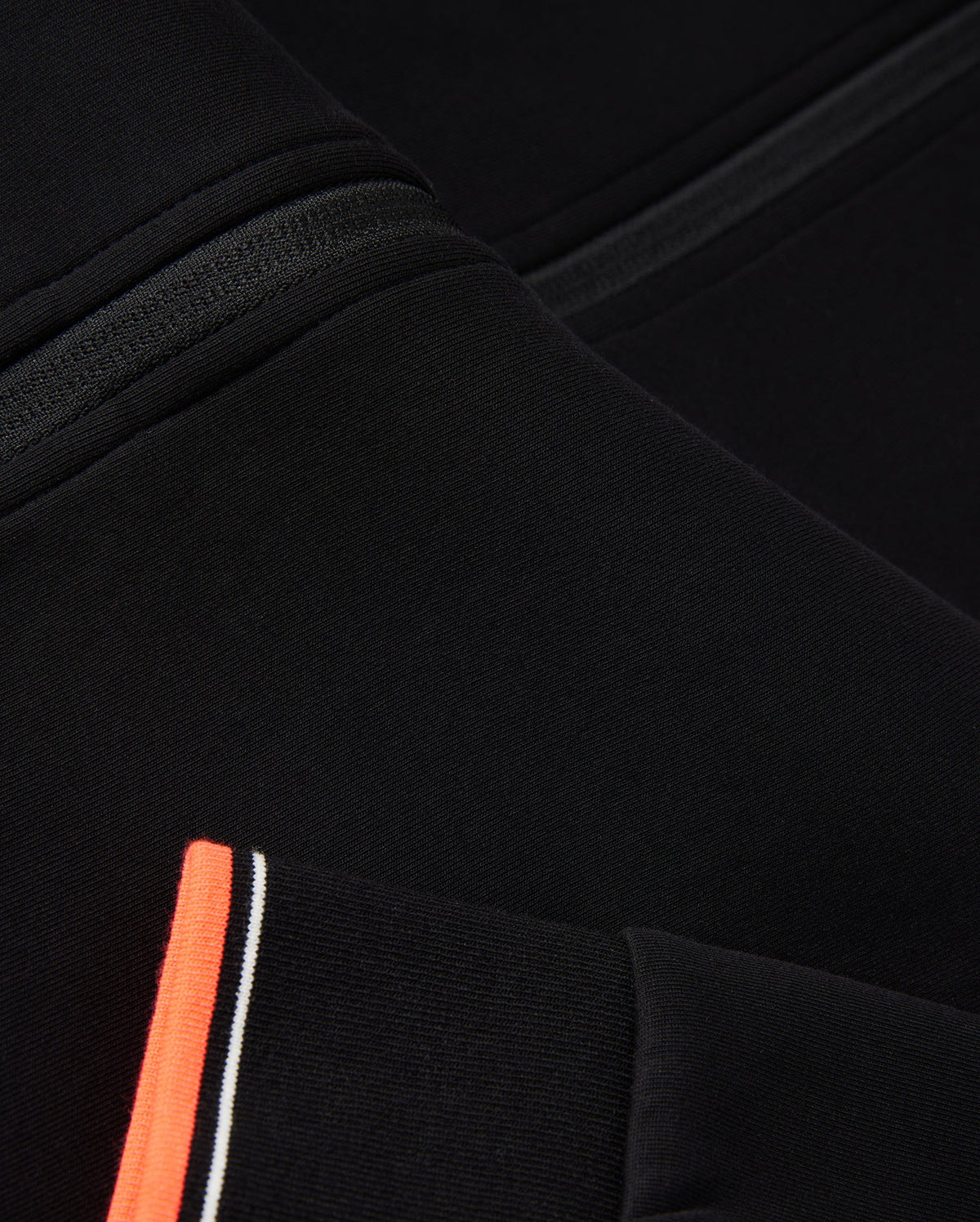 Close-up of a black fabric garment featuring a seam detail and an orange stripe with white and black accents. The image highlights the texture, design elements of the textile, and logo-engraved hardware typical of a Psycho Bunny MENS COURTSIDE TRACK JACKET - B6J692C200.