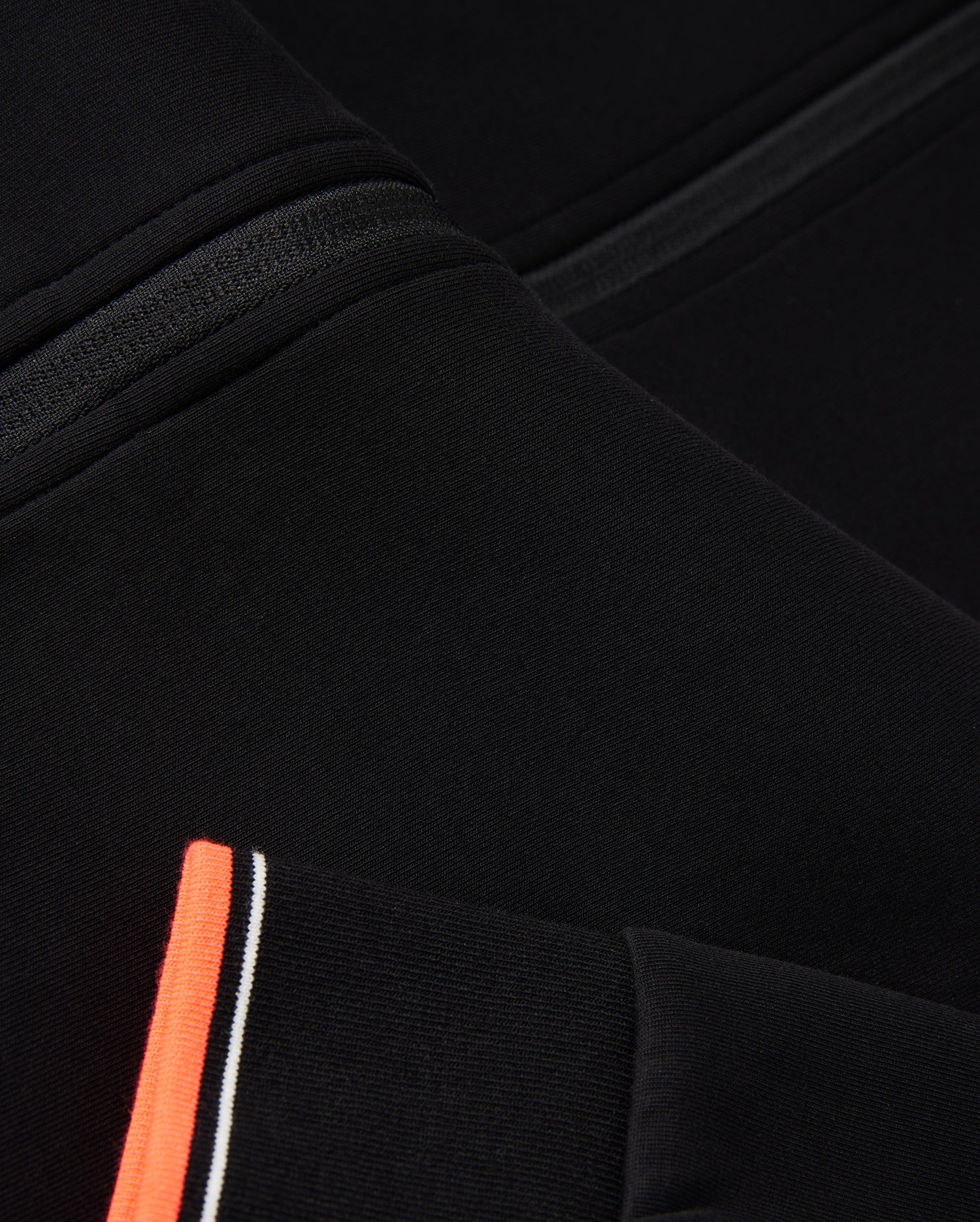 Close-up of a black fabric garment featuring a seam detail and an orange stripe with white and black accents. The image highlights the texture, design elements of the textile, and logo-engraved hardware typical of a Psycho Bunny MENS COURTSIDE TRACK JACKET - B6J692C200.