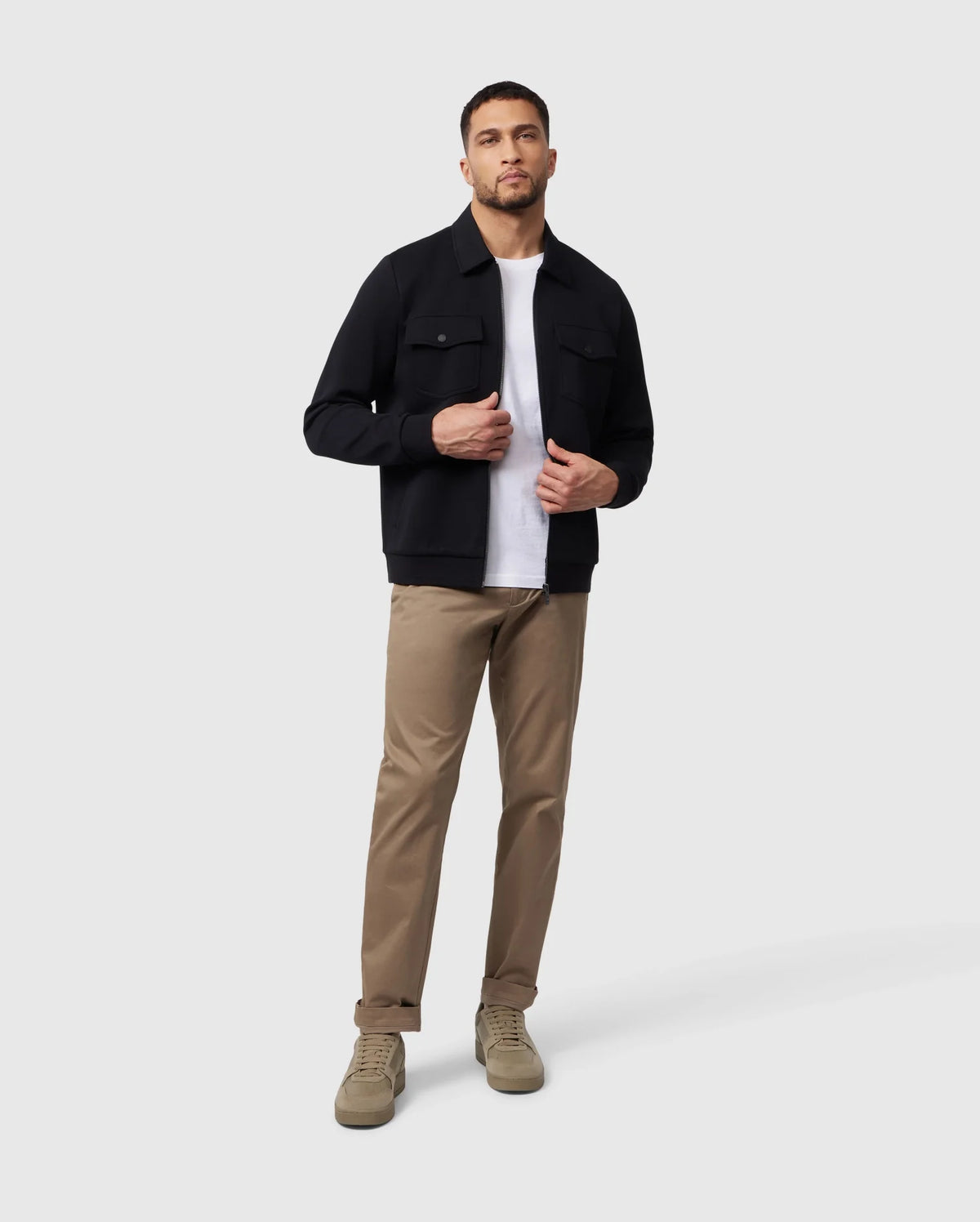 A man stands against a plain light background, donning a stylish MENS OROVILLE BOMBER JACKET - B6J712A200 from Psycho Bunny. It is made of a cotton-poly blend and is paired with a white T-shirt, beige pants, and tan sneakers. He casually has his hands in his pockets while gazing forward.