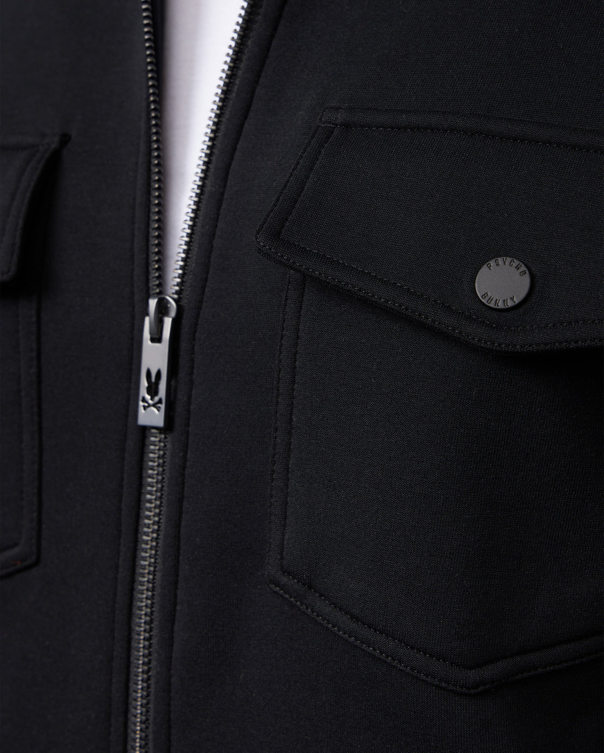 Close-up of the MENS OROVILLE BOMBER JACKET - B6J712A200 by Psycho Bunny, a black bomber jacket featuring a zipper with the brand's distinctive bunny logo on the pull. This outerwear piece showcases two snap-button flap pockets, crafted from a smooth and slightly textured cotton-poly fabric blend.