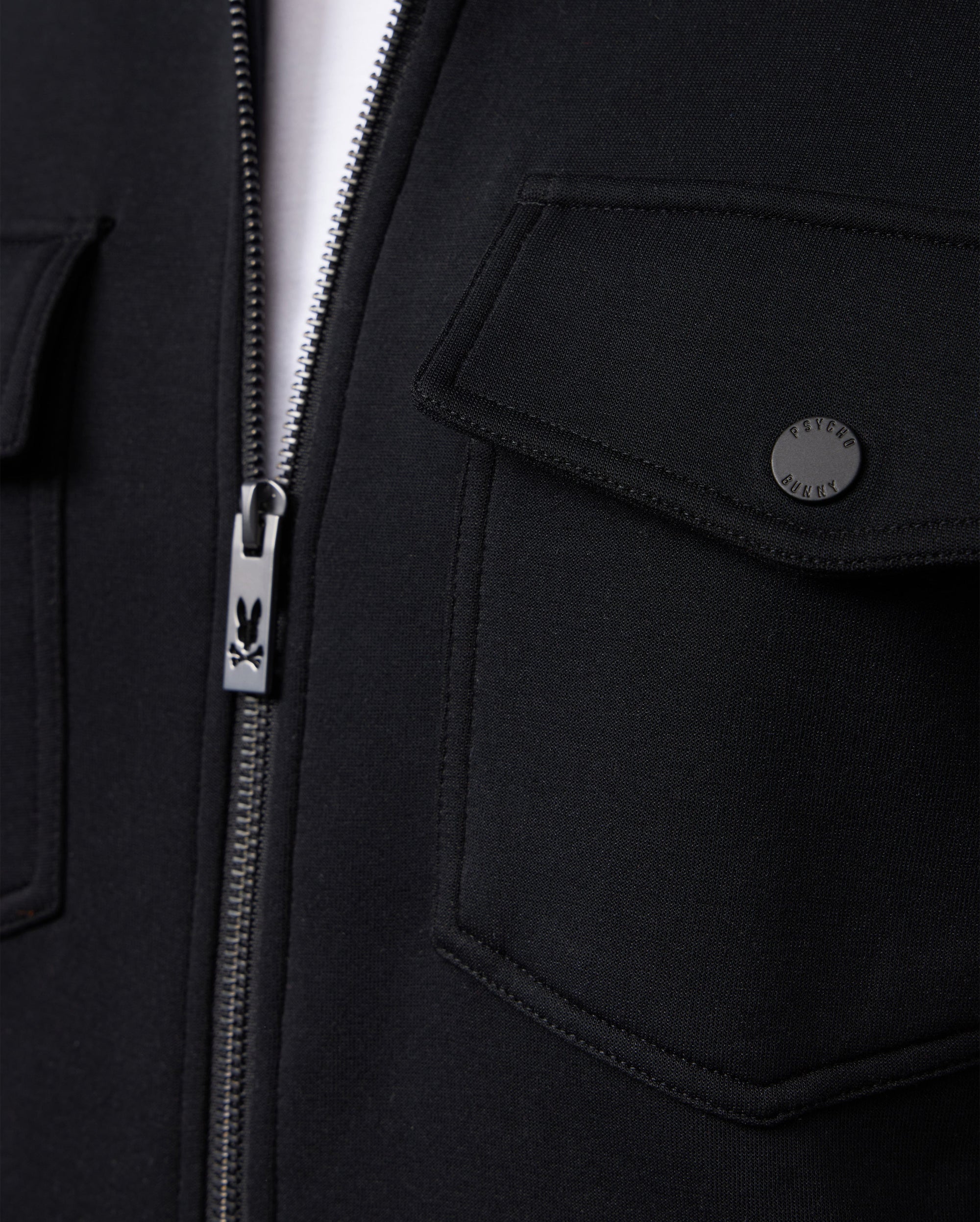 Close-up of the MENS OROVILLE BOMBER JACKET - B6J712A200 by Psycho Bunny, a black bomber jacket featuring a zipper with the brand's distinctive bunny logo on the pull. This outerwear piece showcases two snap-button flap pockets, crafted from a smooth and slightly textured cotton-poly fabric blend.
