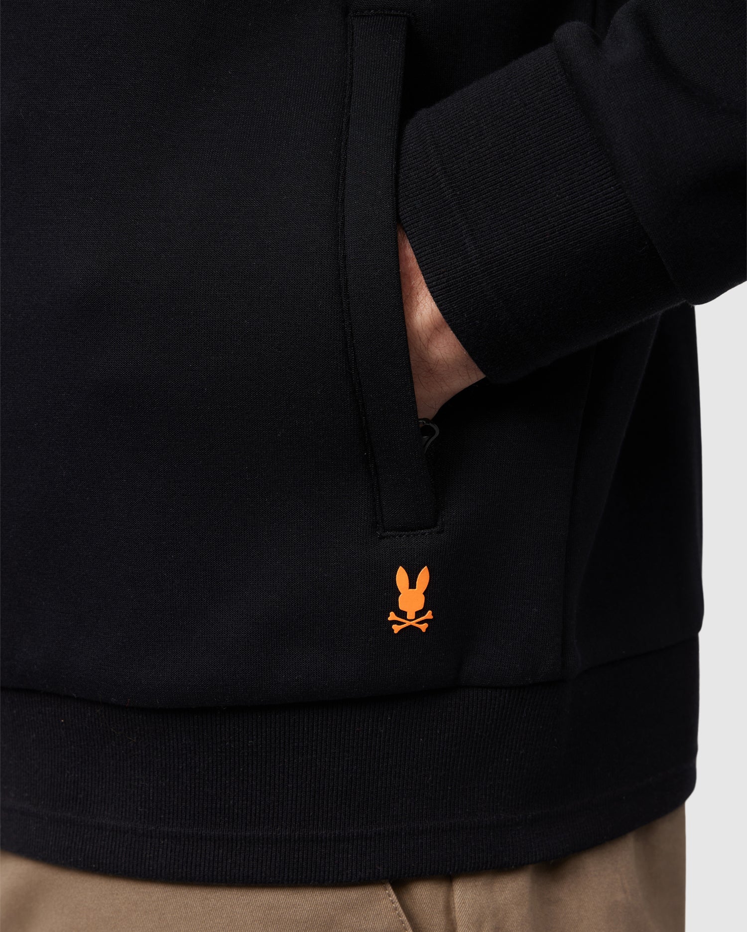 A person is wearing the MENS OROVILLE BOMBER JACKET - B6J712A200 by Psycho Bunny in black, casually tucking one hand into their pocket. This cotton-poly blend jacket stands out with its striking orange bunny with crossbones logo near the hem. They are also styled with beige pants to complete the look.