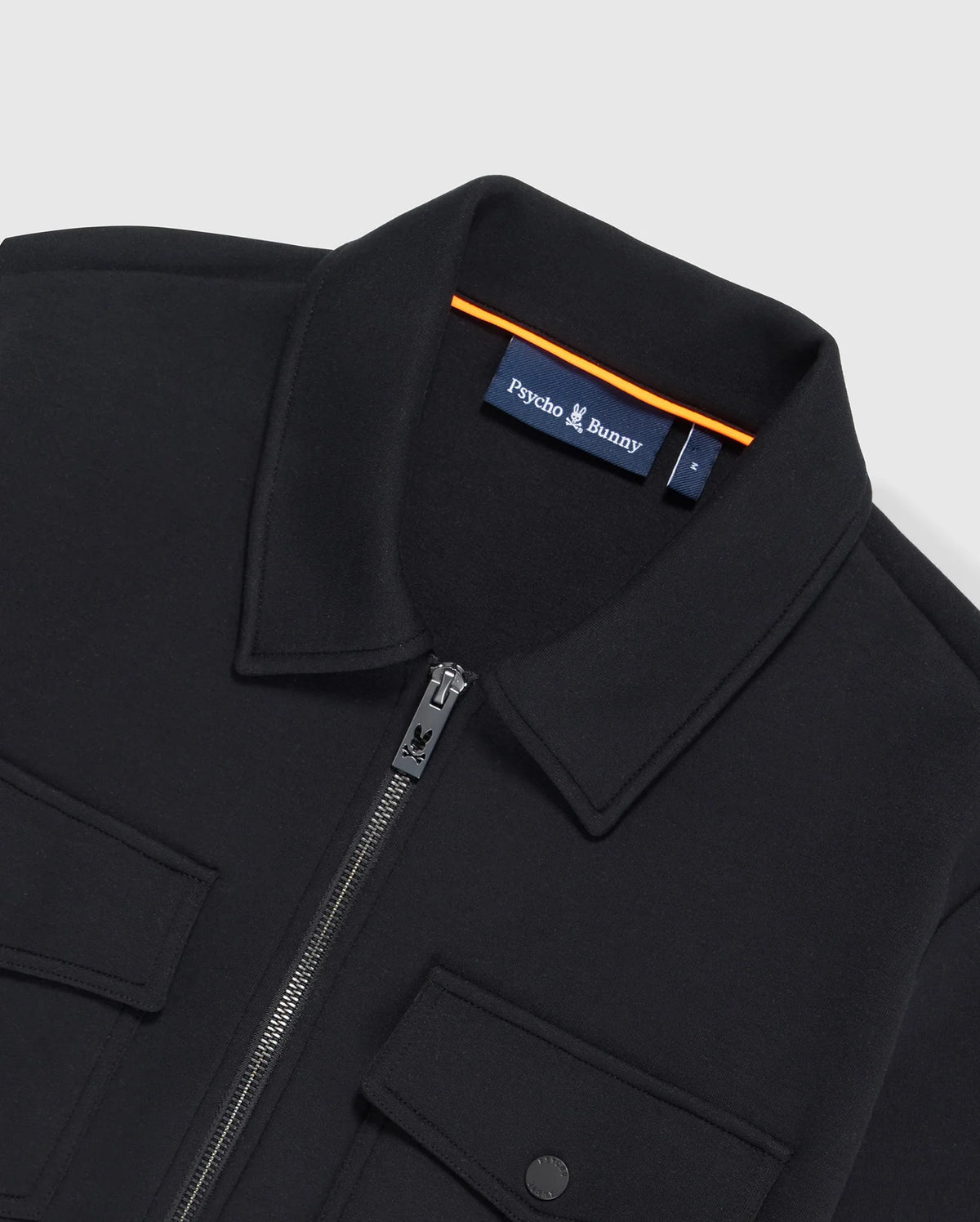 Close-up of the MENS OROVILLE BOMBER JACKET - B6J712A200 in black, featuring a zipper and two chest pockets with button flaps. This stylish piece from the brand "Psycho Bunny" is displayed inside the collar with an eye-catching orange trim accent, and it is crafted from a durable cotton-poly fabric.