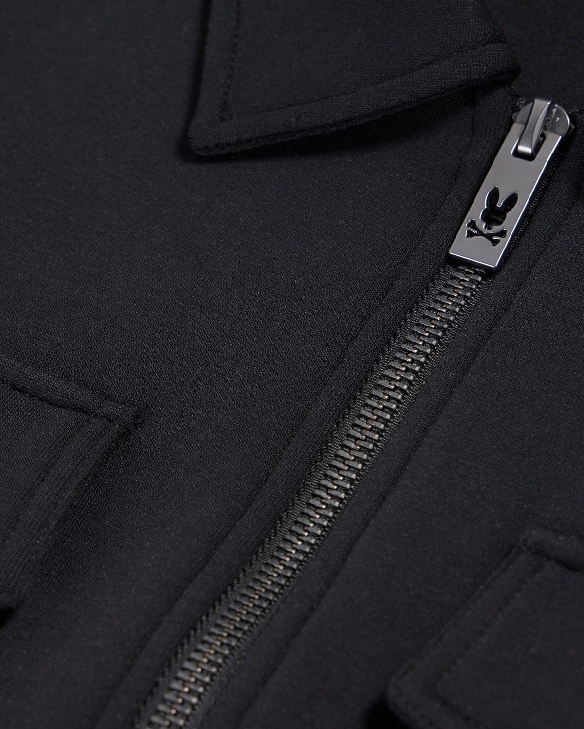 Close-up of the Psycho Bunny MENS OROVILLE BOMBER JACKET - B6J712A200 in black, showcasing a silver zipper with an engraved logo. The smooth cotton-poly fabric and the design featuring folded-over edges hint at a pocket or flap detail near the zipper, enhancing its sleek outerwear appeal.
