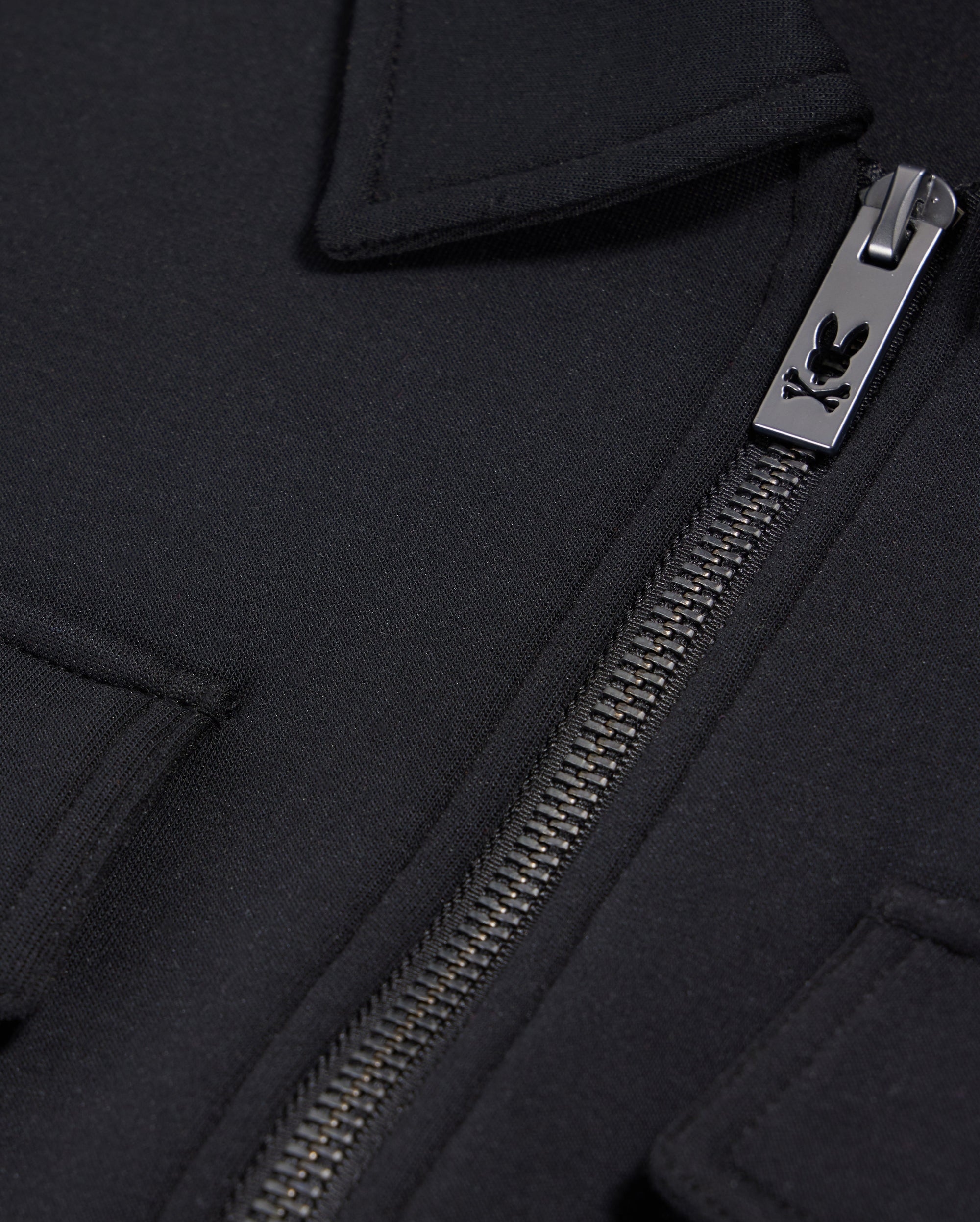 Close-up of the Psycho Bunny MENS OROVILLE BOMBER JACKET - B6J712A200 in black, showcasing a silver zipper with an engraved logo. The smooth cotton-poly fabric and the design featuring folded-over edges hint at a pocket or flap detail near the zipper, enhancing its sleek outerwear appeal.