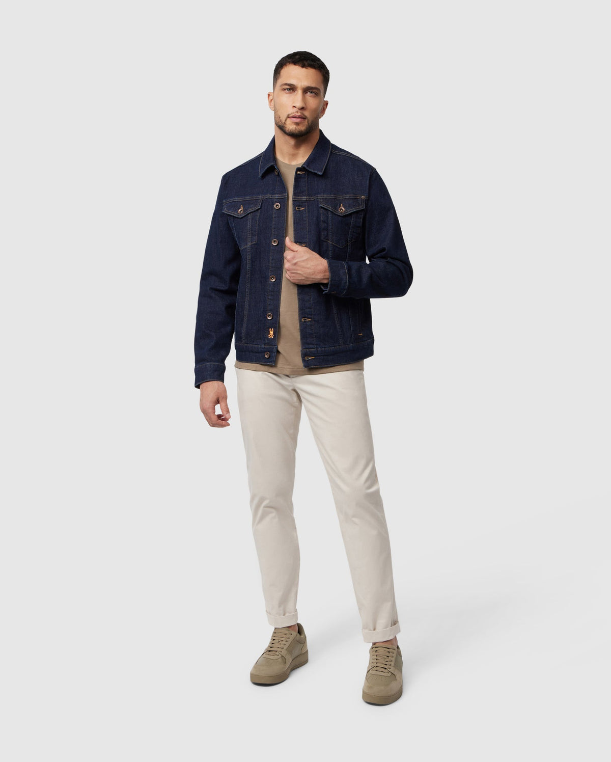 A man stands against a light gray background wearing the Psycho Bunny MENS PATHE DENIM JACKET - B6J741X1CO, beige pants, and olive green shoes. He has short hair and a trimmed beard, with his right hand holding the edge of his jacket.