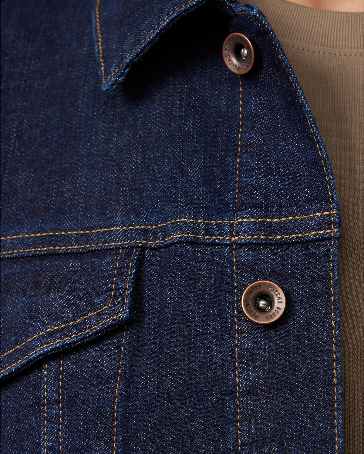 Close-up of the MENS PATHE DENIM JACKET - B6J741X1CO by Psycho Bunny, showcasing detailed stitching and copper buttons, complete with an adjustable fit against a brown shirt background.