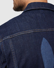 A man is wearing the Psycho Bunny MENS PATHE DENIM JACKET - B6J741X1CO, which offers an adjustable fit. The image highlights a subtle vertical design detail on the back of the jacket. The man's face isn't shown, and he stands against a plain background.