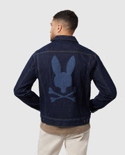 A man sports the Psycho Bunny MENS PATHE DENIM JACKET - B6J741X1CO, featuring an embroidered bunny skull and crossbones design on the back. With his back to the camera, he effortlessly pairs it with a brown shirt and beige pants against a plain background, exuding coolness.