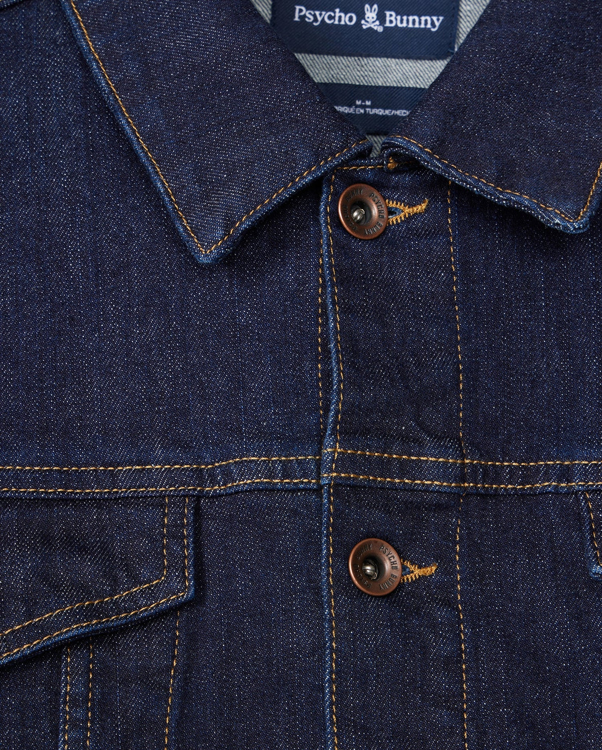 Close-up of the MENS PATHE DENIM JACKET - B6J741X1CO, showcasing its collar and two-button design. The label at the neck reads "Psycho Bunny." Visible stitching adds detail to the rugged fabric, making it a standout piece in men's fashion.
