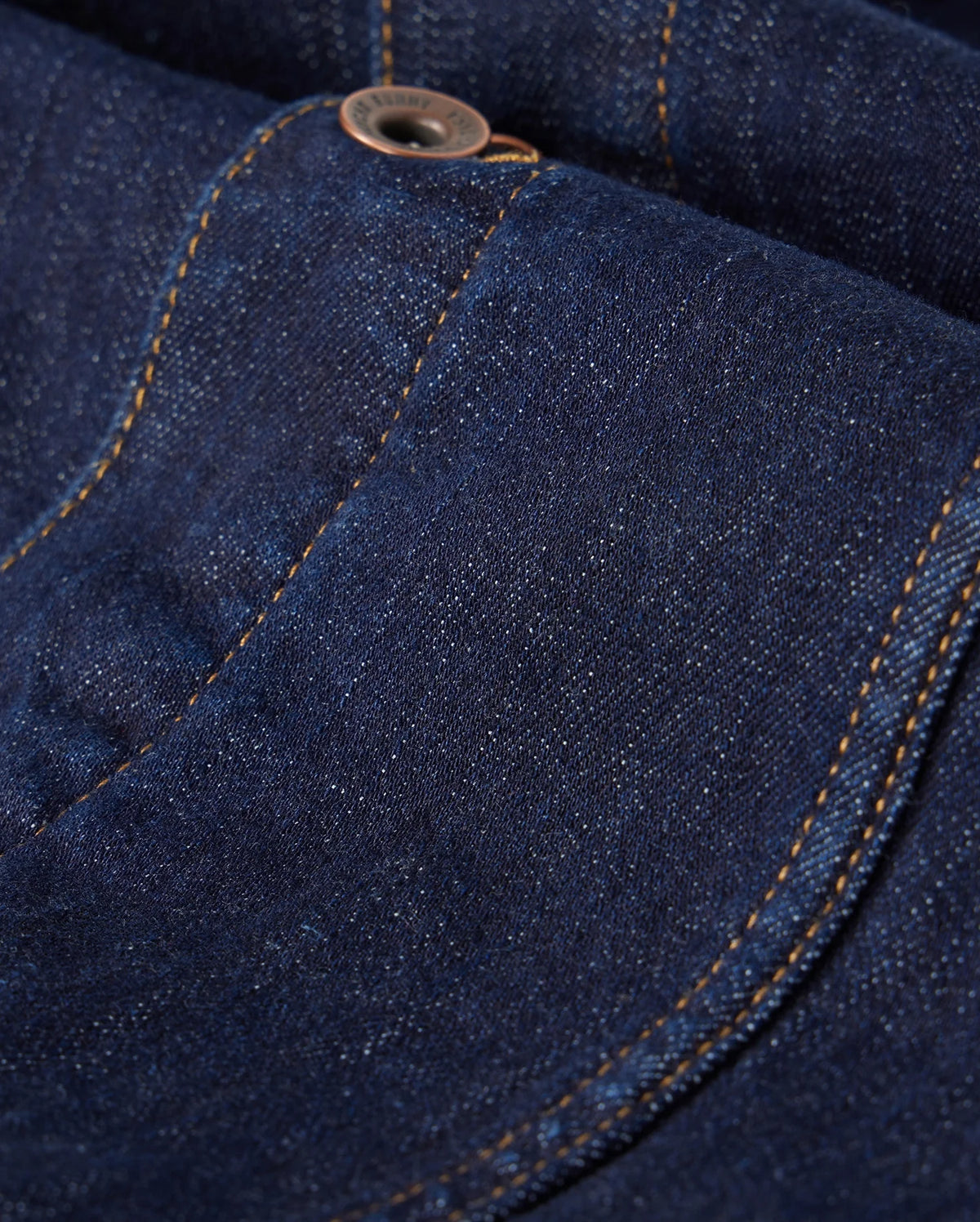 The MENS PATHE DENIM JACKET - B6J741X1CO by Psycho Bunny features a close-up view of dark blue denim with an apparent pocket and button. The orange stitching contrasts with the fabric, and embroidered details enhance the texture, creating a striking appearance.
