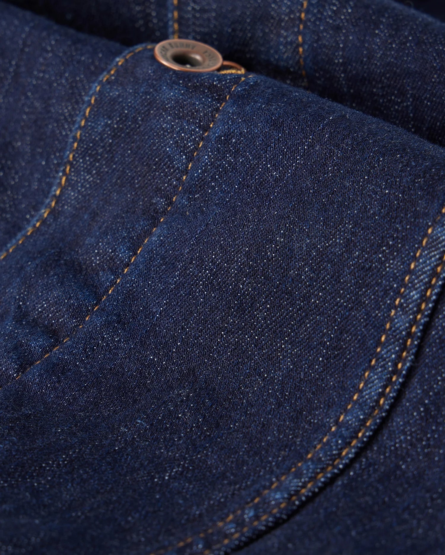 The MENS PATHE DENIM JACKET - B6J741X1CO by Psycho Bunny features a close-up view of dark blue denim with an apparent pocket and button. The orange stitching contrasts with the fabric, and embroidered details enhance the texture, creating a striking appearance.