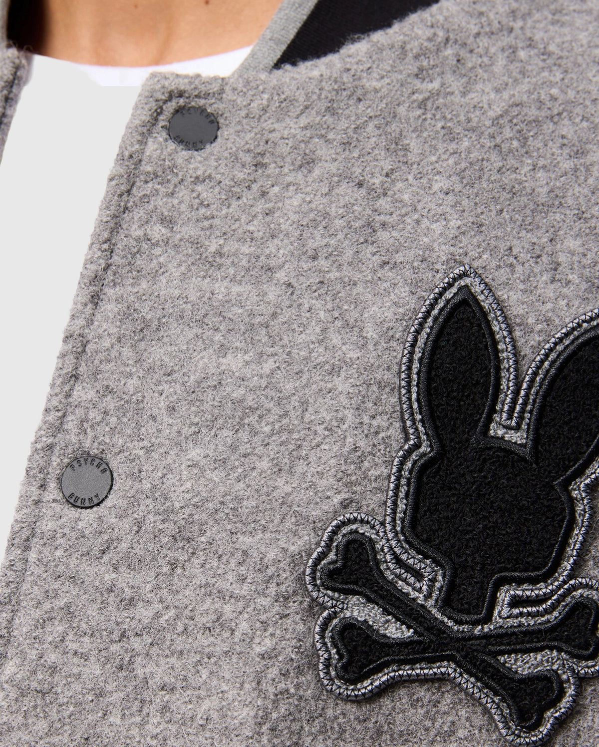 Close-up of the stylish Men's Tom Wool Bomber Jacket by Psycho Bunny, featuring a gray design adorned with a black rabbit and crossbones patch on the side. The jacket, identified as product B6J744D200, showcases a round black button with part of a white shirt peeking out from beneath.