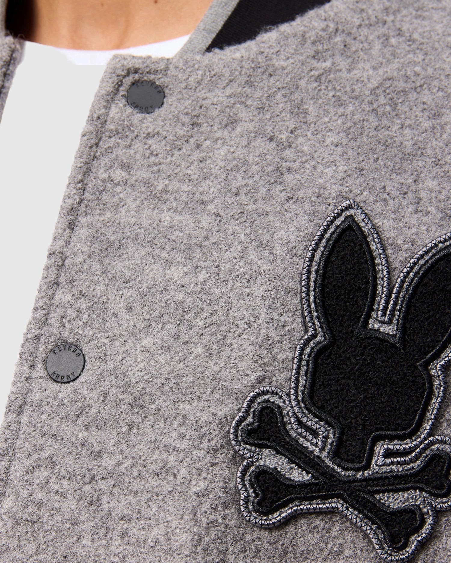 Close-up of the stylish Men's Tom Wool Bomber Jacket by Psycho Bunny, featuring a gray design adorned with a black rabbit and crossbones patch on the side. The jacket, identified as product B6J744D200, showcases a round black button with part of a white shirt peeking out from beneath.
