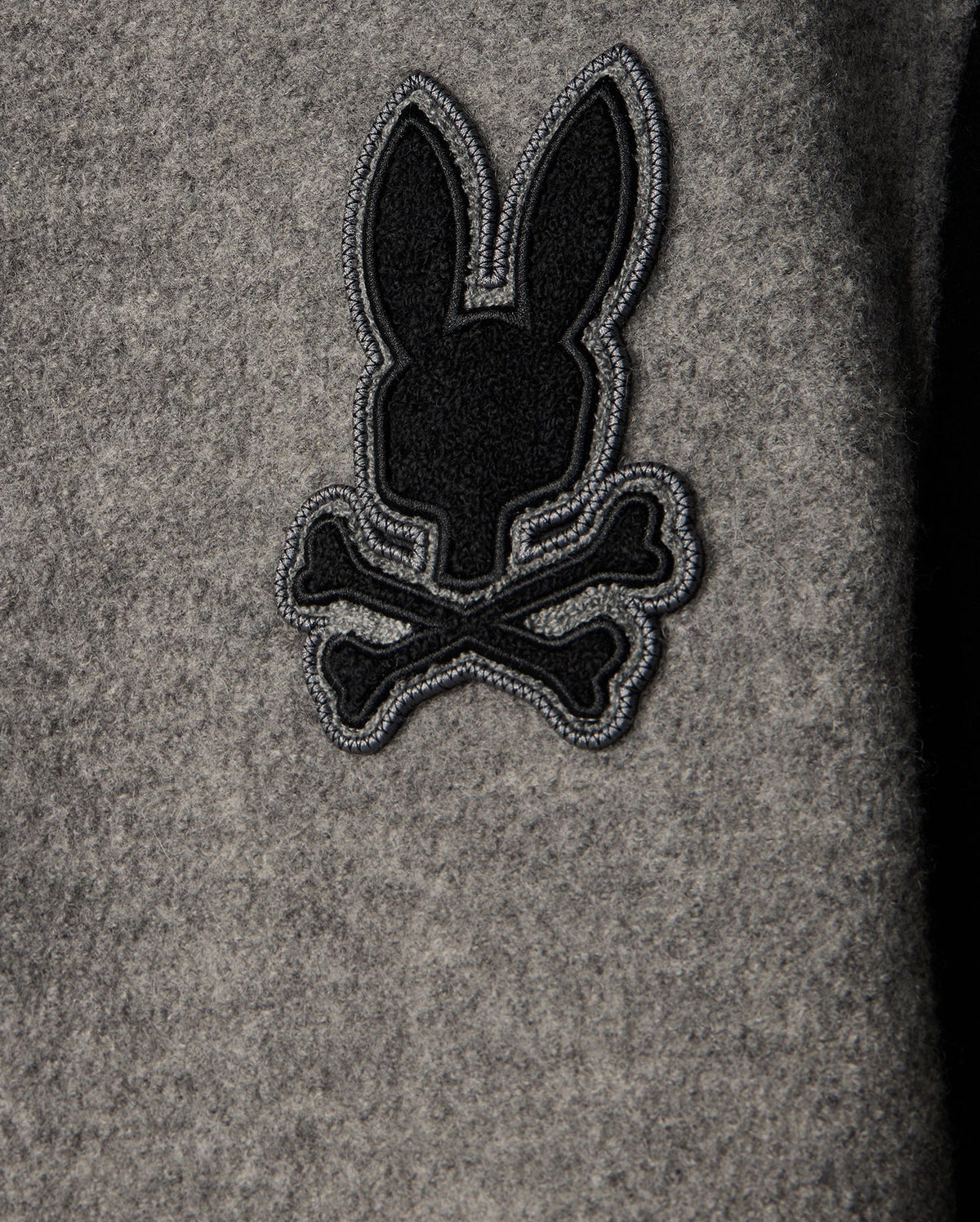The MENS TOM WOOL BOMBER JACKET - B6J744D200 by Psycho Bunny showcases a stylish gray wool design with a playful black embroidery of a rabbit silhouette above crossed bones, offering an elegant twist to the classic skull-and-crossbones motif. Ideal for those who appreciate distinctive details in men's fashion.