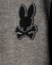 The MENS TOM WOOL BOMBER JACKET - B6J744D200 by Psycho Bunny showcases a stylish gray wool design with a playful black embroidery of a rabbit silhouette above crossed bones, offering an elegant twist to the classic skull-and-crossbones motif. Ideal for those who appreciate distinctive details in men's fashion.