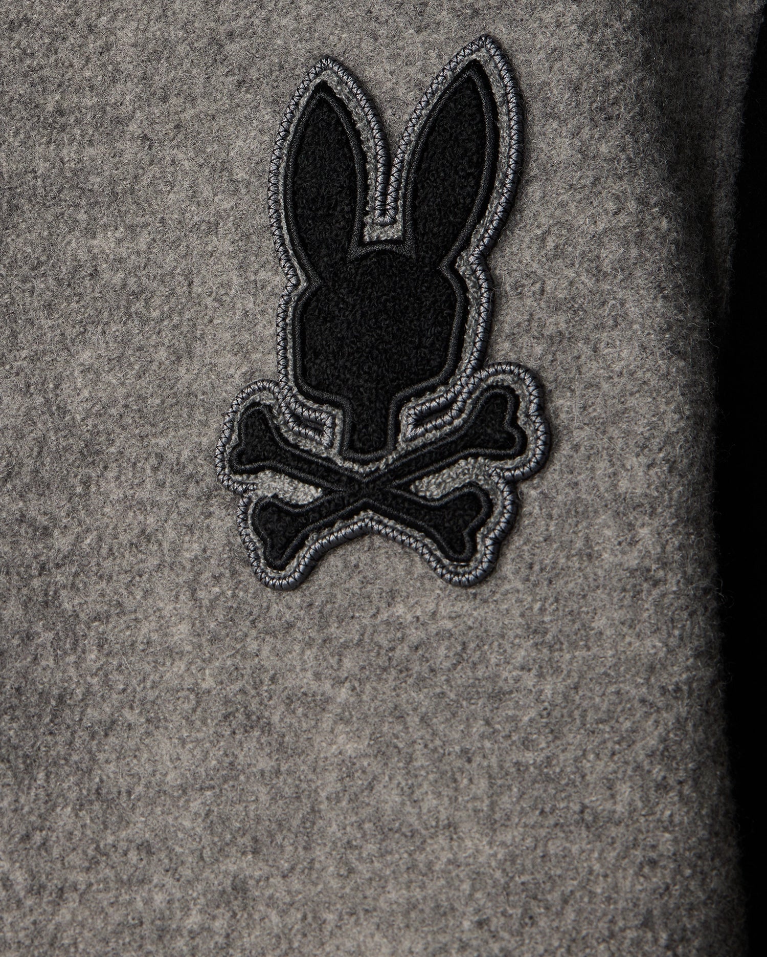 The MENS TOM WOOL BOMBER JACKET - B6J744D200 by Psycho Bunny showcases a stylish gray wool design with a playful black embroidery of a rabbit silhouette above crossed bones, offering an elegant twist to the classic skull-and-crossbones motif. Ideal for those who appreciate distinctive details in men's fashion.