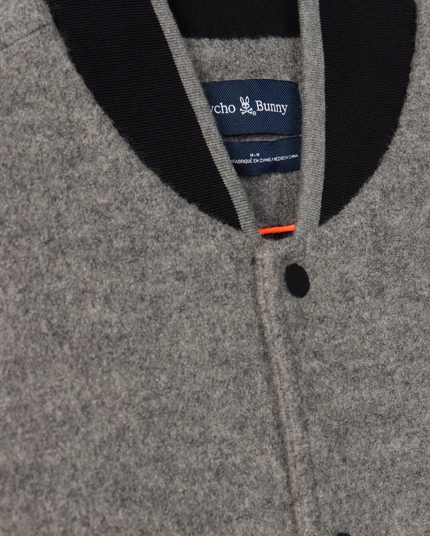Close-up of the MENS TOM WOOL BOMBER JACKET - B6J744D200 by Psycho Bunny in grey, featuring a black collar and black buttons. The elegant touch is enhanced by a blue label with the Psycho Bunny logo visible on the inside.