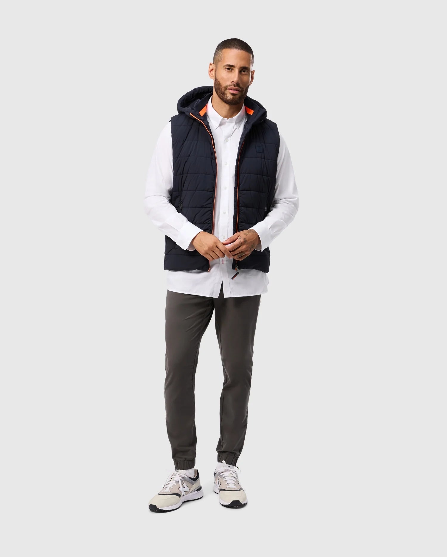 A man stands against a plain background, wearing a Psycho Bunny MENS MALCOLM MIXED MEDIA VEST (B6J861D200), a white button-up shirt, olive green pants, and white sneakers. He has short hair and a beard, with his hands clasped in front.