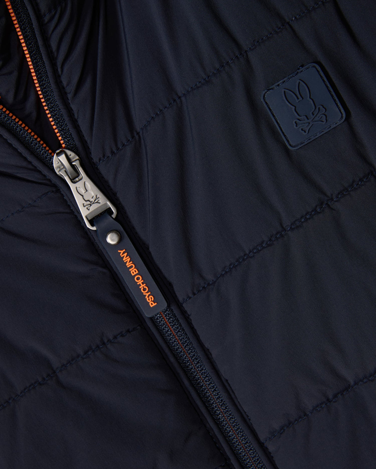 Close-up of the Psycho Bunny Men's Malcolm Mixed Media Vest (B6J861D200) in black, featuring a metal zipper with an orange pull tab reading "Psycho Bunny" and a small embroidered bunny logo on a fabric patch.