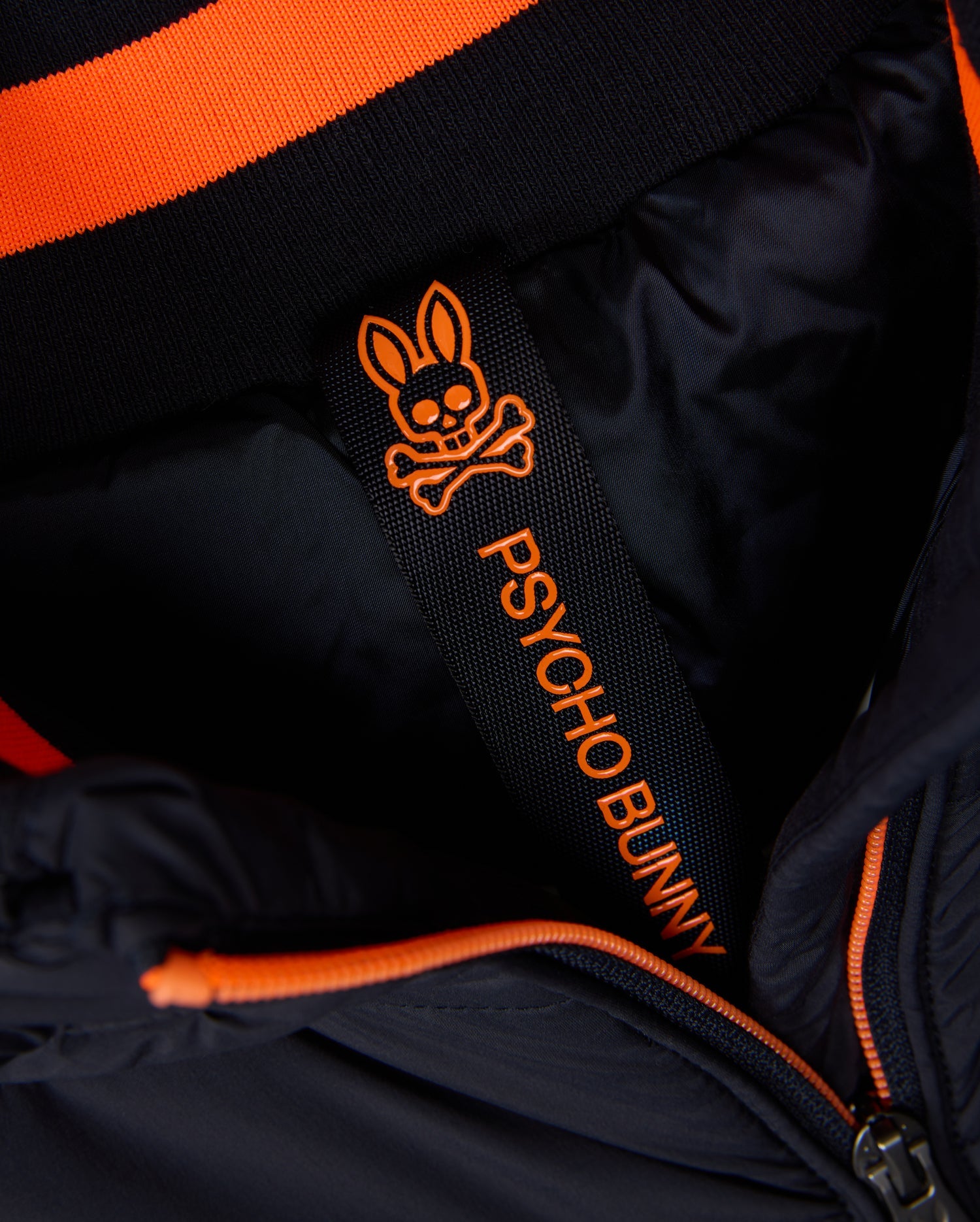 The MENS MALCOLM MIXED MEDIA VEST - B6J861D200 by Psycho Bunny features a black and orange padded design. Inside, a label showcases the brand’s signature skull and bunny logo. The collar is highlighted with striking orange and black stripes.