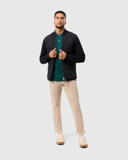 A man stands confidently in the MENS MYKEL MIXED MEDIA JACKET by Psycho Bunny, a versatile black piece worn over a green polo shirt. The outfit is completed with beige pants and white sneakers, set against a plain light gray background that highlights his casual yet stylish attire.
