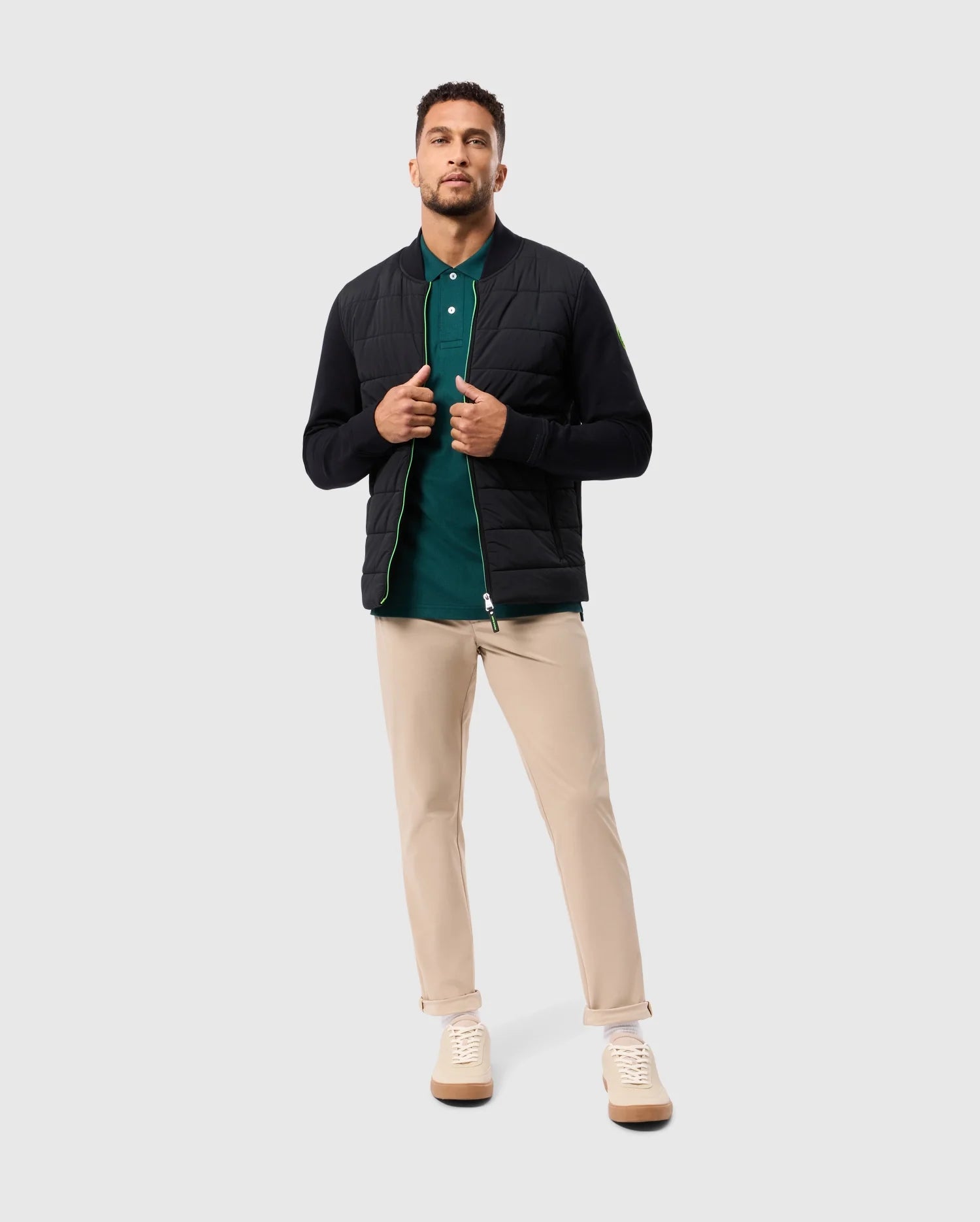 A man stands confidently in the MENS MYKEL MIXED MEDIA JACKET by Psycho Bunny, a versatile black piece worn over a green polo shirt. The outfit is completed with beige pants and white sneakers, set against a plain light gray background that highlights his casual yet stylish attire.