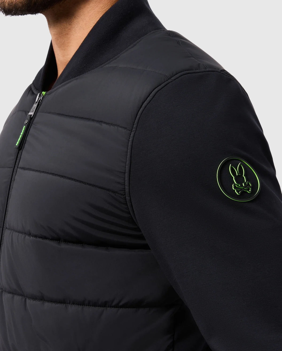 A person wearing the MENS MYKEL MIXED MEDIA JACKET - B6J875D200 by Psycho Bunny, a versatile black padded design with a green-lined rabbit emblem on the sleeve. This men's jacket, featuring a zipper and contrasting plain background, combines style and practicality. Only the upper body is visible.