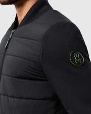 A person wearing the MENS MYKEL MIXED MEDIA JACKET - B6J875D200 by Psycho Bunny, a versatile black padded design with a green-lined rabbit emblem on the sleeve. This men's jacket, featuring a zipper and contrasting plain background, combines style and practicality. Only the upper body is visible.